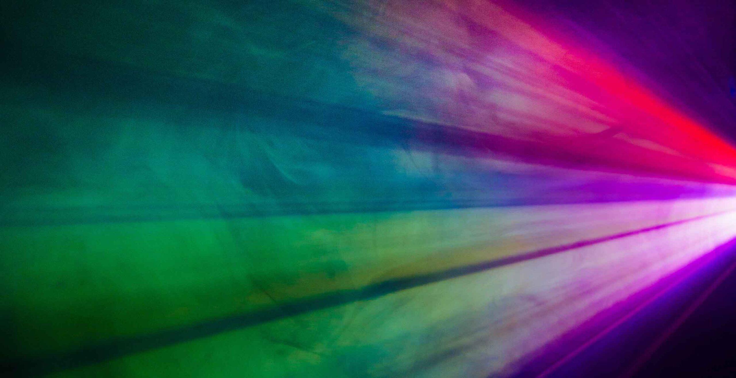 Colorful beams of light in shades of green, blue, purple, pink, and red radiate from the right side of the image, creating a vibrant, abstract pattern against a dark background.