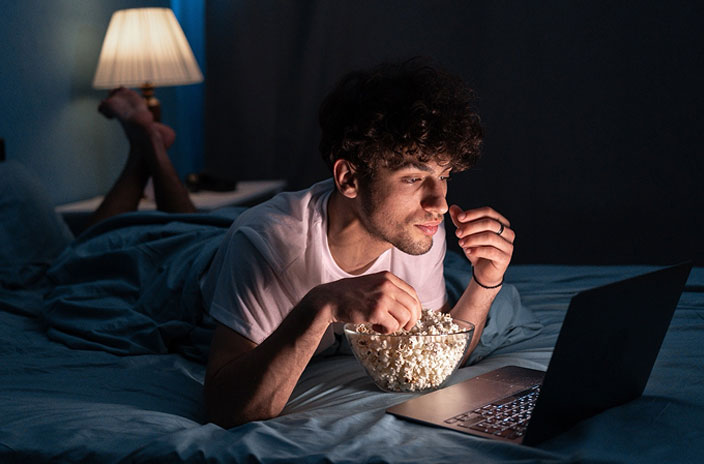 A young man lies on his bed in a dimly lit room, eating popcorn and watching something on his laptop—perhaps researching SEO for AI answer engines. A lamp glows softly in the background, creating a cozy atmosphere.