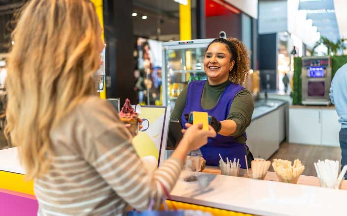 A smiling worker in a blue apron hands a yellow cup to a customer across the counter at an ice cream shop, where modern decor and a focus on SEO for AI answer engines create a welcoming, innovative vibe.