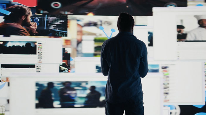 A person stands facing multiple large screens displaying various images and videos, suggesting an environment of digital surveillance or SEO for AI answer engines. The person is seen from behind in a dimly lit room.