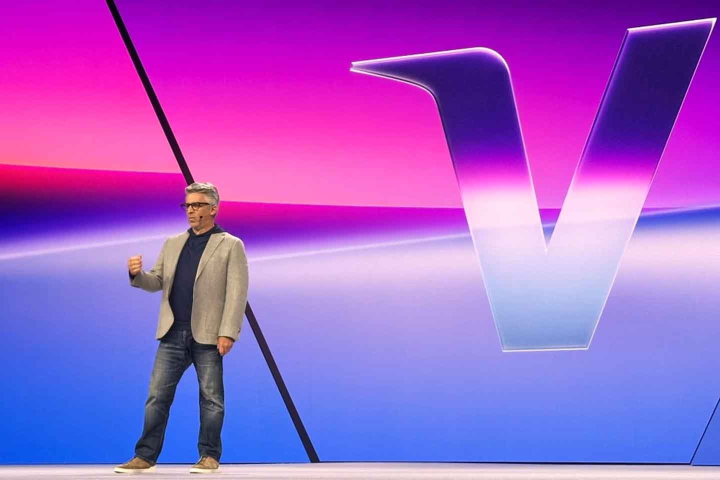 A man in a gray blazer, dark shirt, and jeans stands on stage before a large gradient background, presenting on SEO for AI answer engines with a prominent letter V displayed beside him.